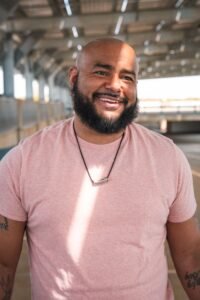 A bald, bearded man smiling warmly while standing in a train station.